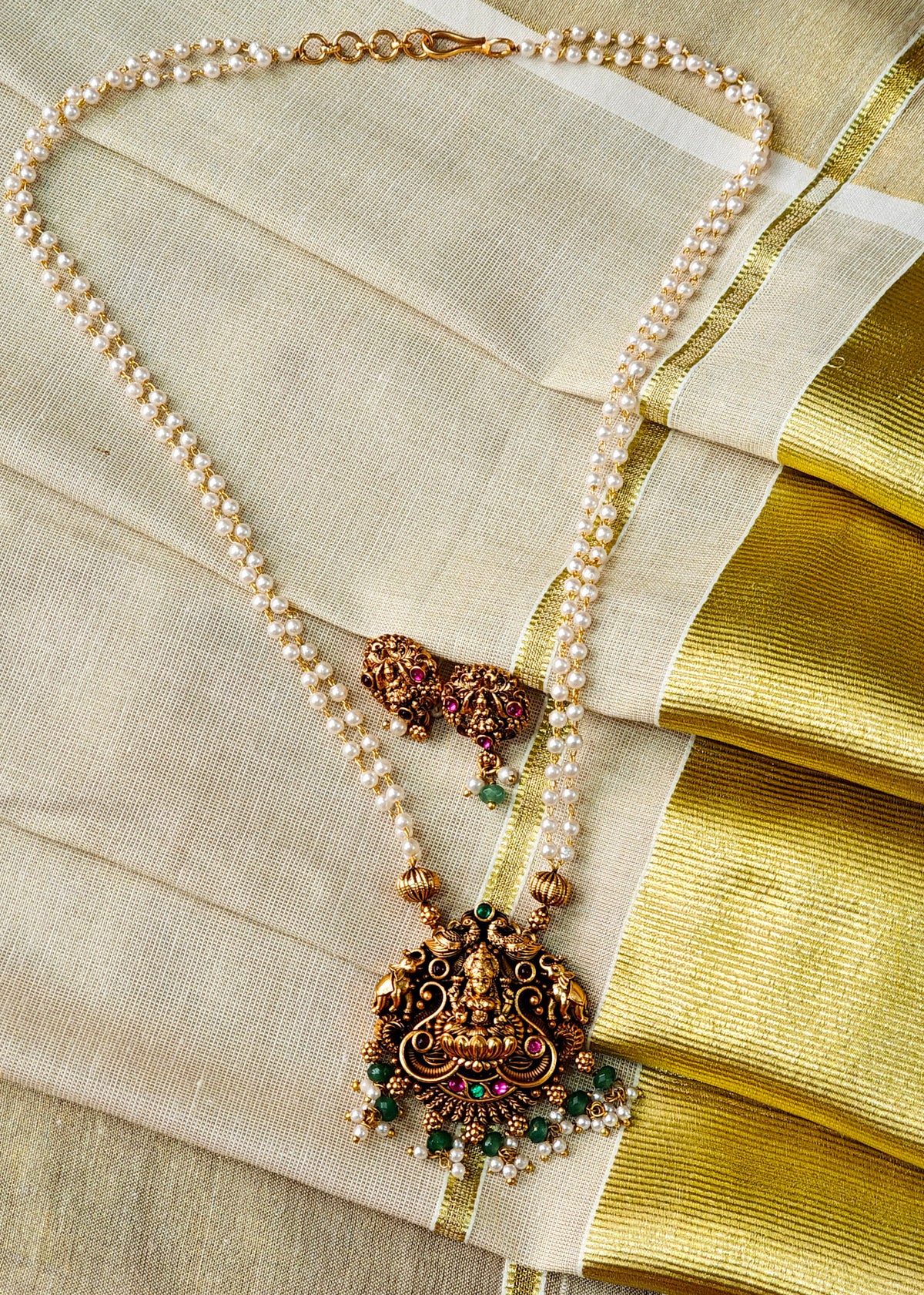 An ornate traditional necklace with a large gold-toned Lakshmi pendant, decorated with green and red stones and small pearl-like beads, displayed on a cream and gold sari fabric.