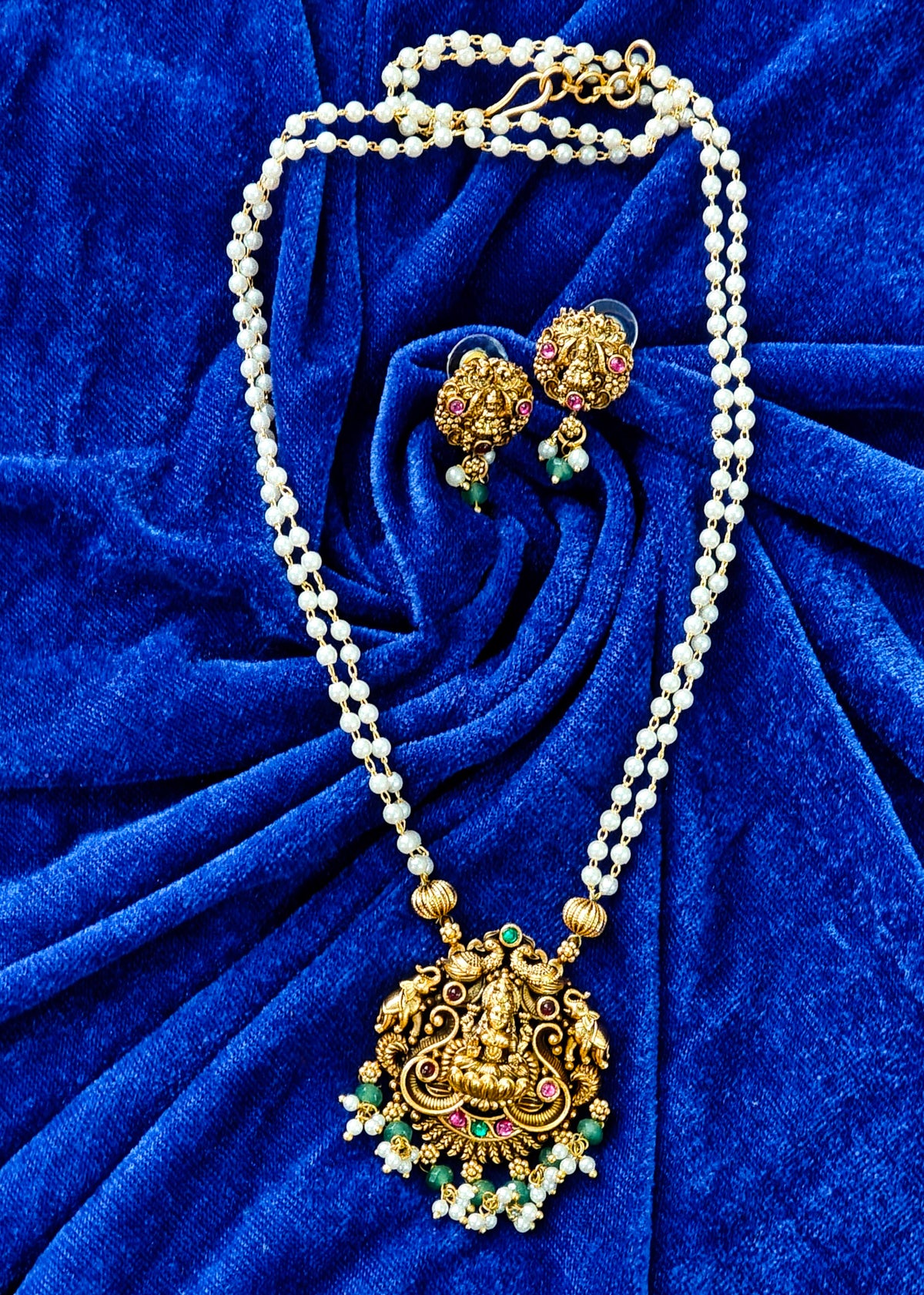 Gold and white pearl jewelry set featuring a traditional pendant necklace and matching earrings, displayed on a textured royal blue velvet background.