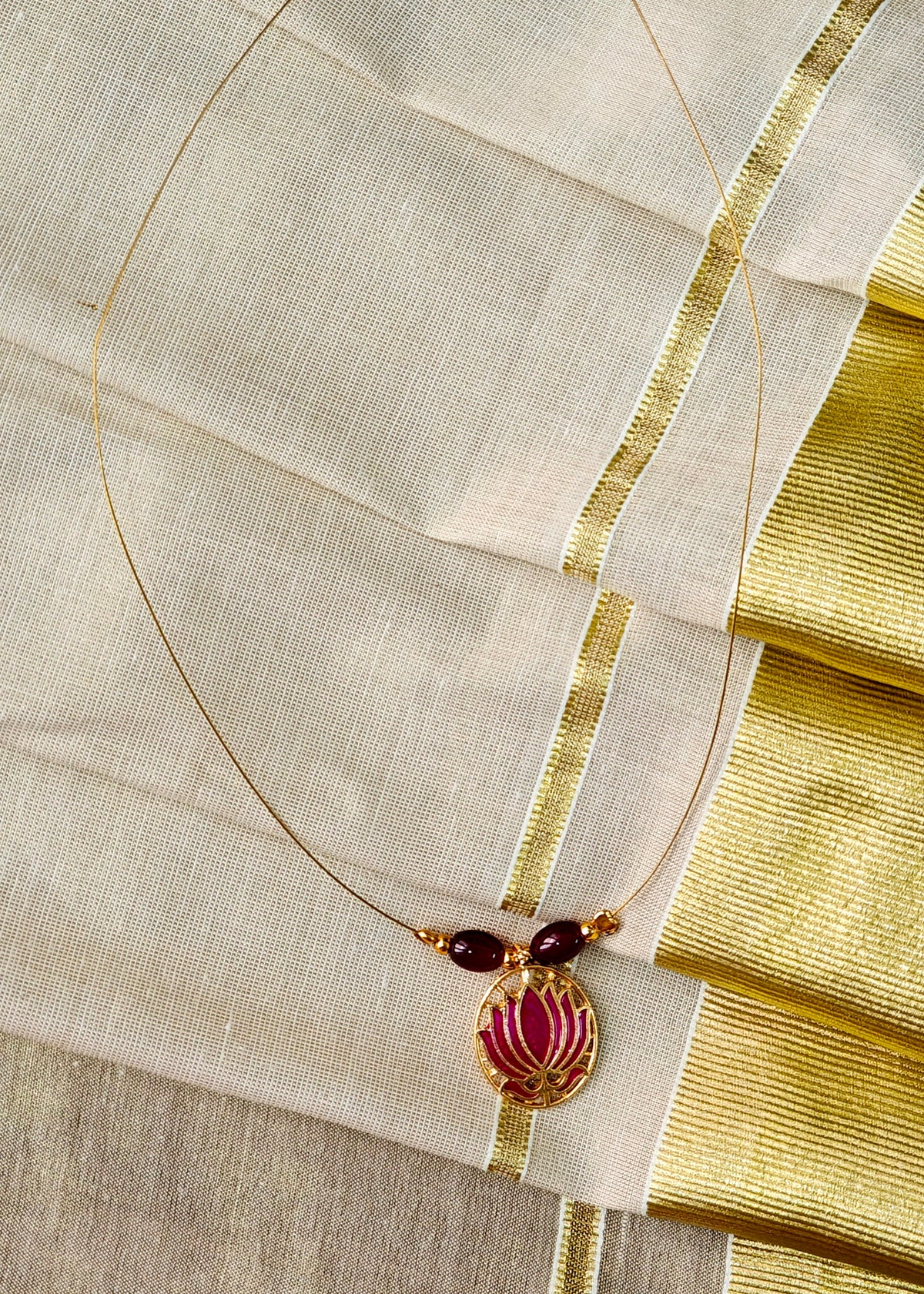 A gold-toned necklace with two dark red beads and a central gold pendant featuring a pink floral motif, displayed on a cream-colored fabric with gold borders.