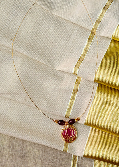 A gold-toned necklace with two dark red beads and a central gold pendant featuring a pink floral motif, displayed on a cream-colored fabric with gold borders.