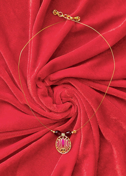 A delicate gold necklace with a circular pendant featuring pink and gold floral details, accented by dark red beads, displayed on a vibrant red fabric background.