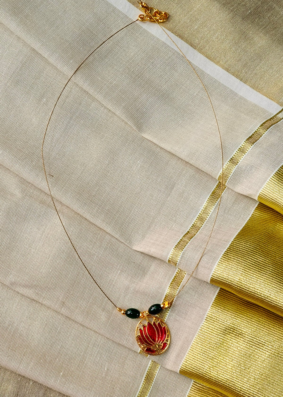 A delicate gold necklace with a lotus pendant, accented by two small black beads, displayed on a draped cream-colored fabric with gold borders.