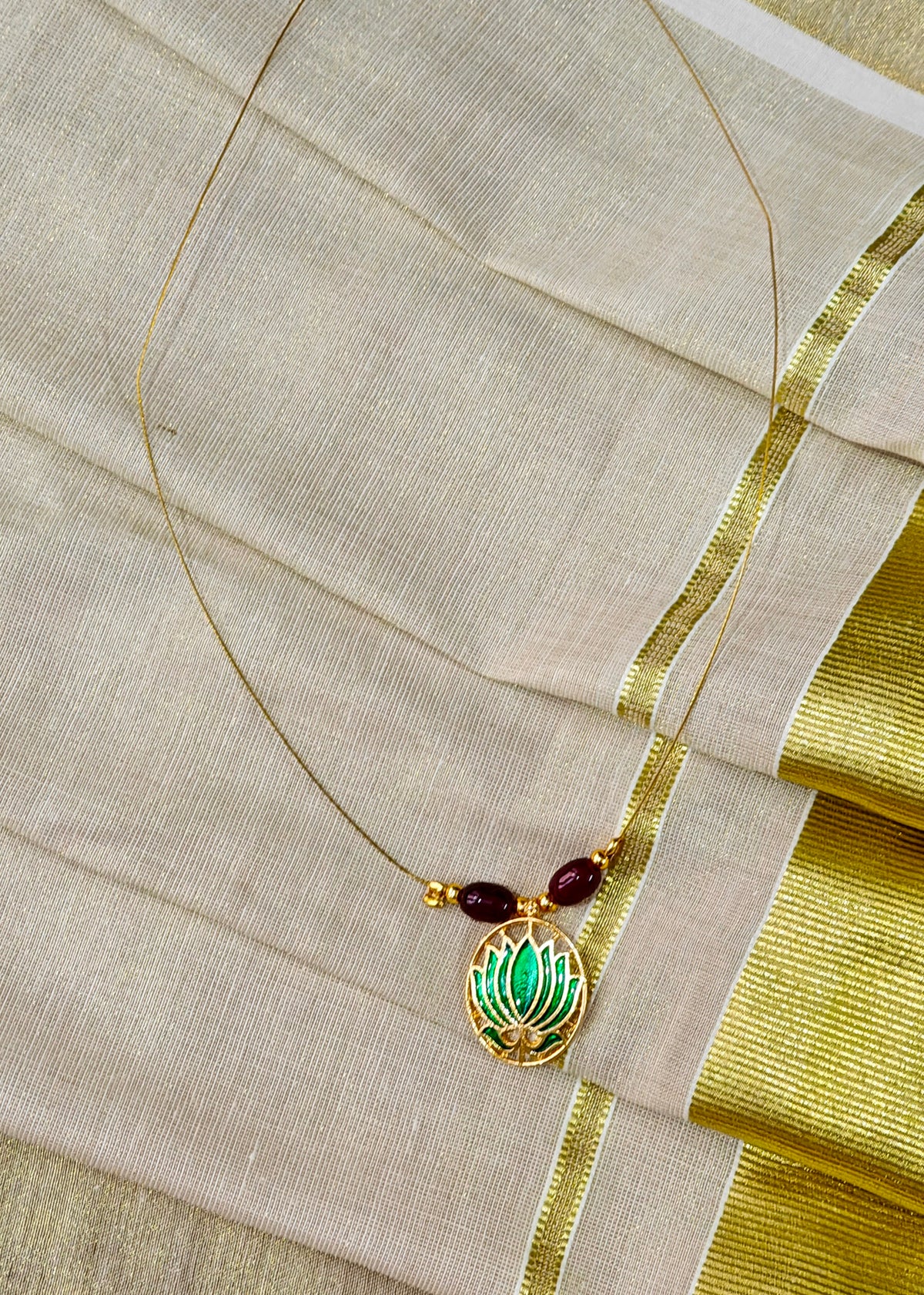 A gold-toned necklace with a lotus-shaped pendant featuring green enamel detailing, laid over a beige fabric with a gold border.