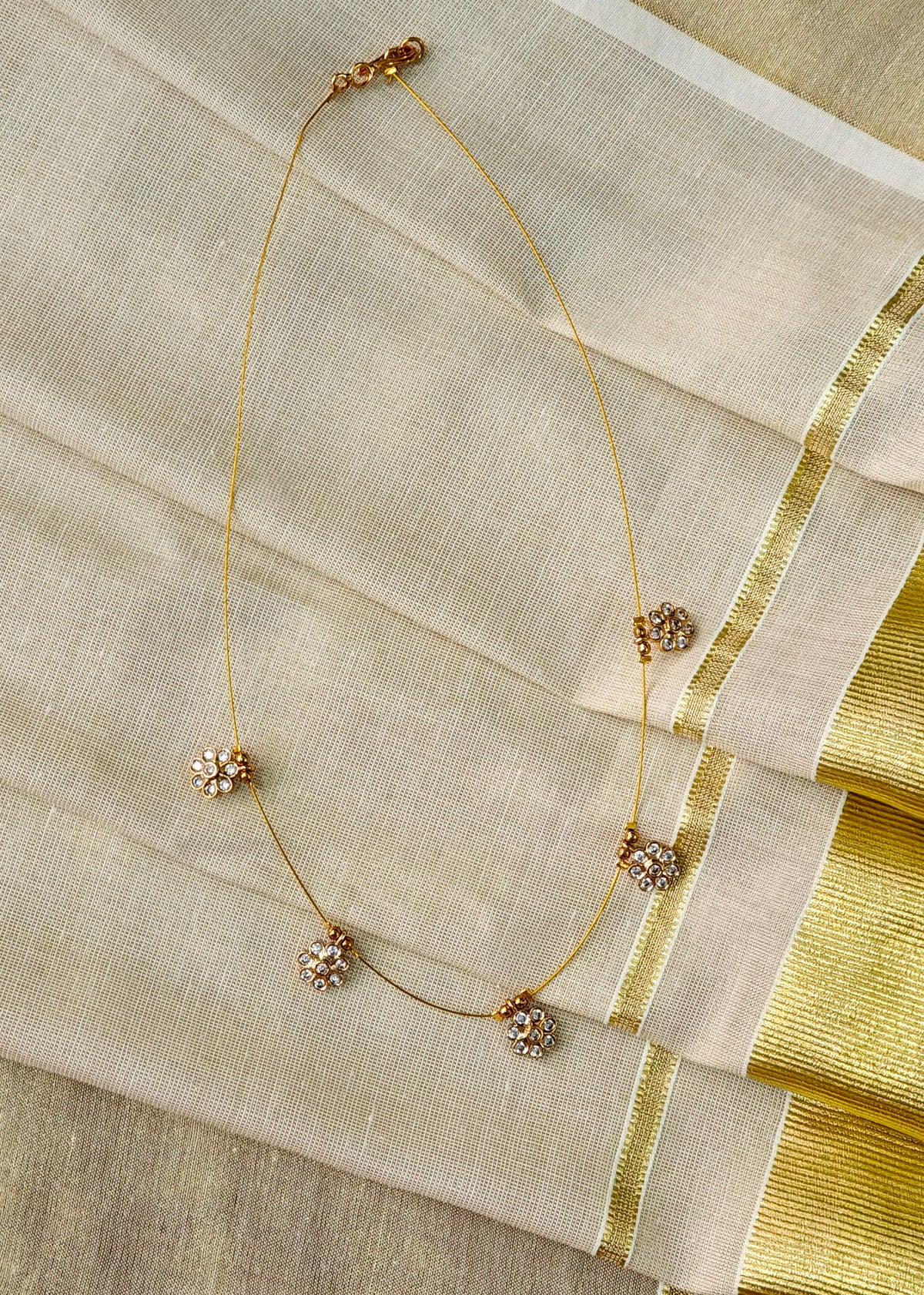 A delicate gold chain necklace with five ornate floral-shaped gold and crystal embellishments, draped elegantly on a folded cream-colored fabric with a gold border.
