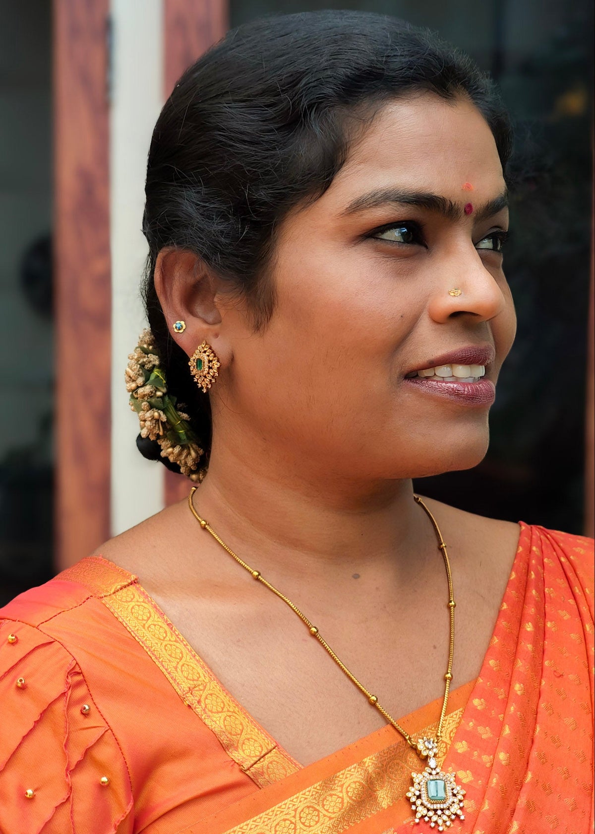 A woman wearing a , a gold necklace with a green stone pendant, and gold earrings, shown in a close-up portrait.