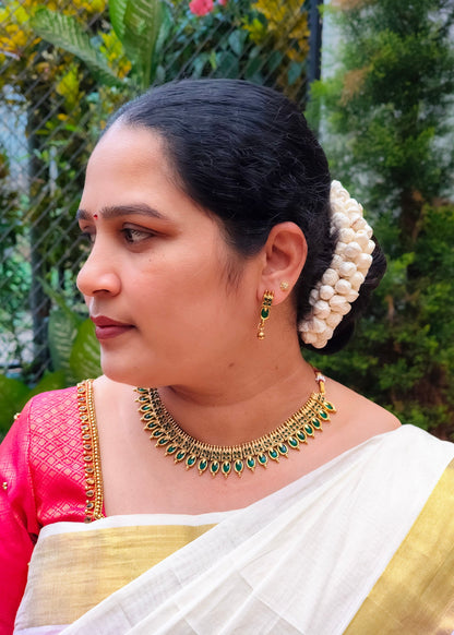 A woman in profile wearing a traditional Indian outfit with a white and gold saree, a pink blouse, a gold necklace with green stones, and a pearl hair accessory, set against a blurred green garden background.
