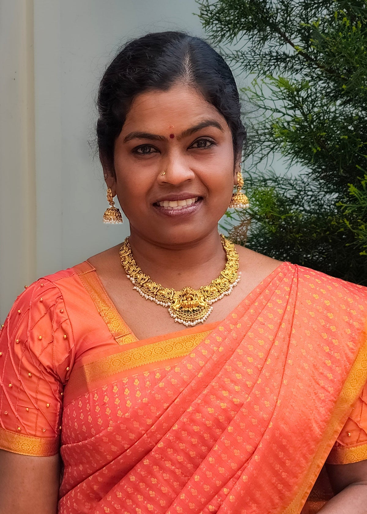 A woman wearing a peach-orange saree with gold patterns and a gold border, paired with a matching embroidered blouse, a gold necklace, and earrings, standing outdoors.