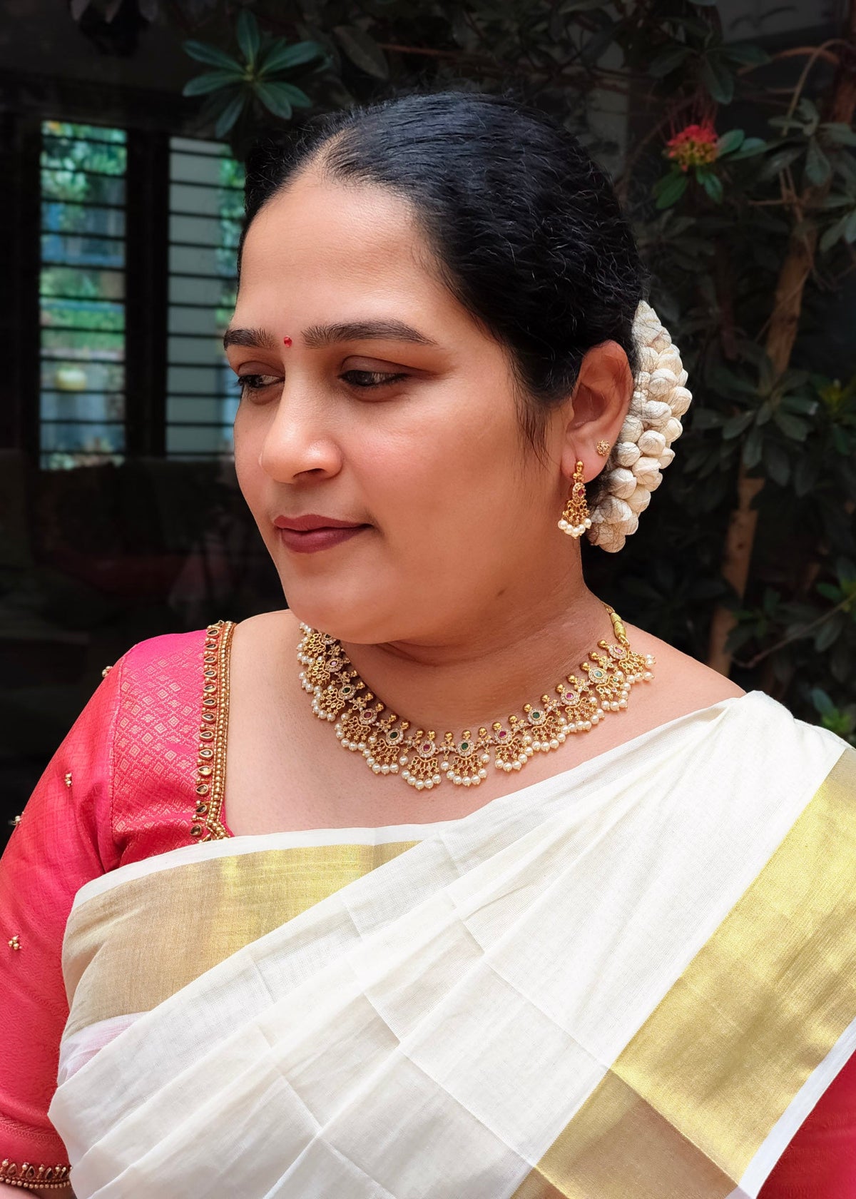 A woman wearing a white and gold saree with a pink blouse, gold jewelry, and a white floral hair accessory, photographed outdoors with greenery in the background.