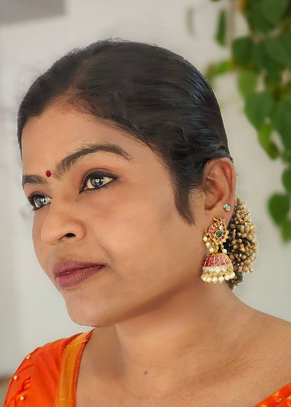 A close-up portrait of a woman wearing large ornate gold-toned jhumka earrings with red and white bead detailing.