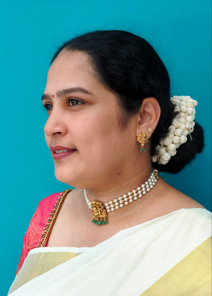 A woman in profile wearing traditional Indian jewelry, including a pearl necklace with a gold pendant, matching earrings, and a white sari with a pink and gold border, set against a solid turquoise blue background.