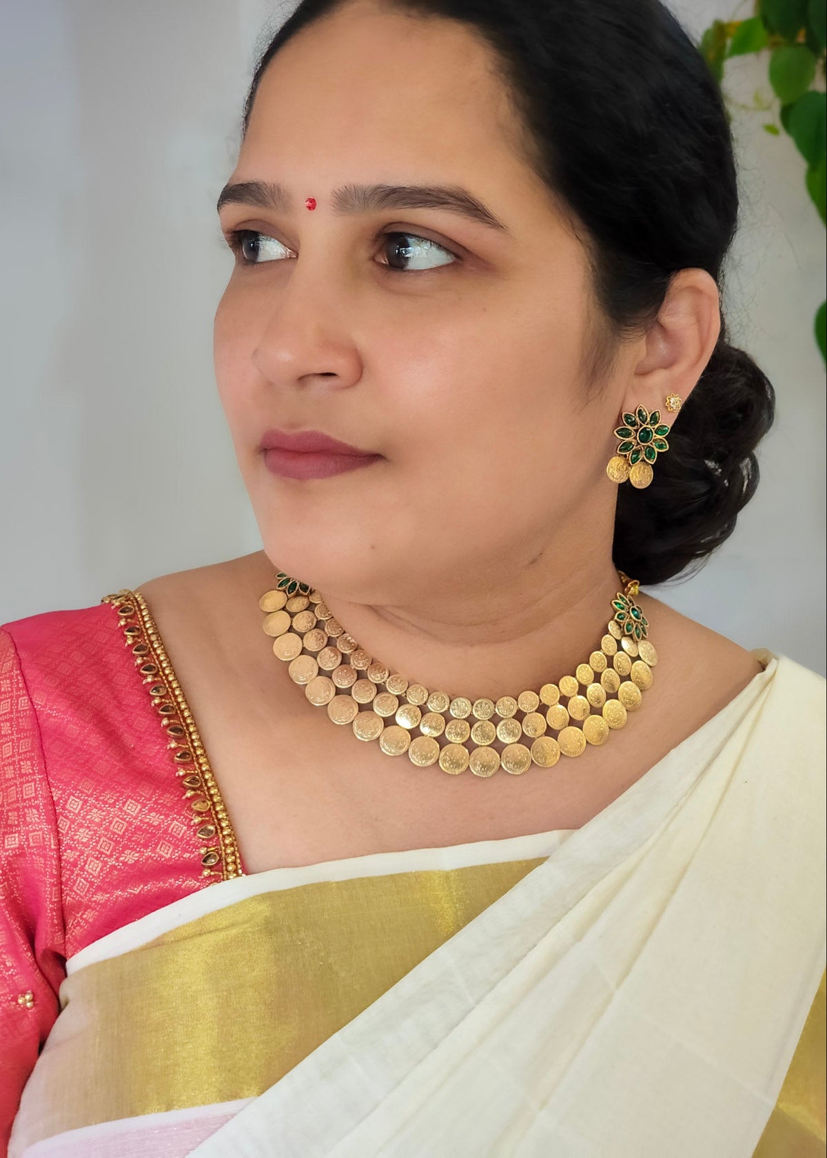 A woman wearing a traditional Indian outfit with a pink and gold embroidered blouse and a white saree, accessorized with a gold coin necklace and matching earrings.