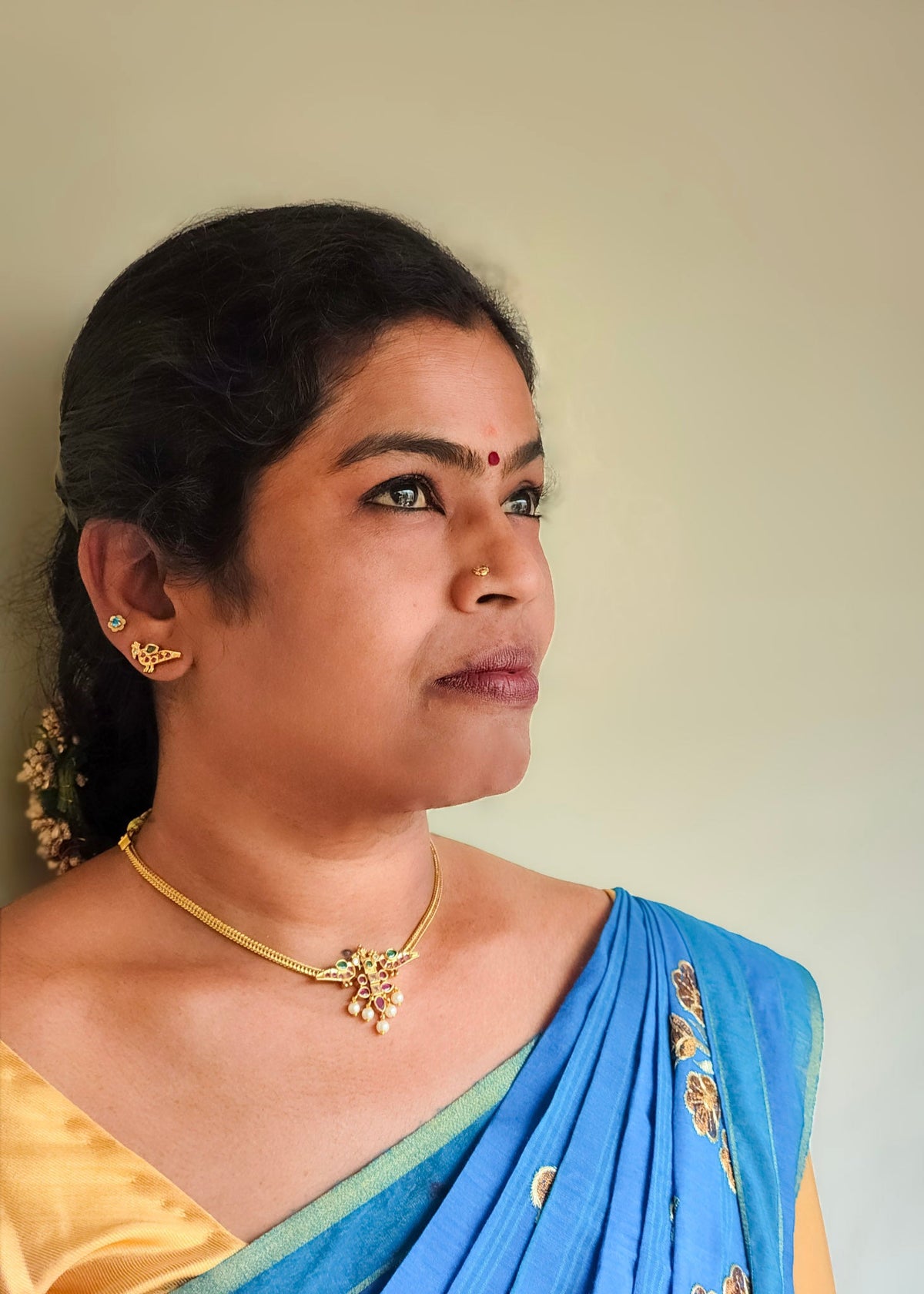 A woman in profile wearing a blue saree with gold embroidery, a yellow blouse, and a gold necklace against a plain beige wall.