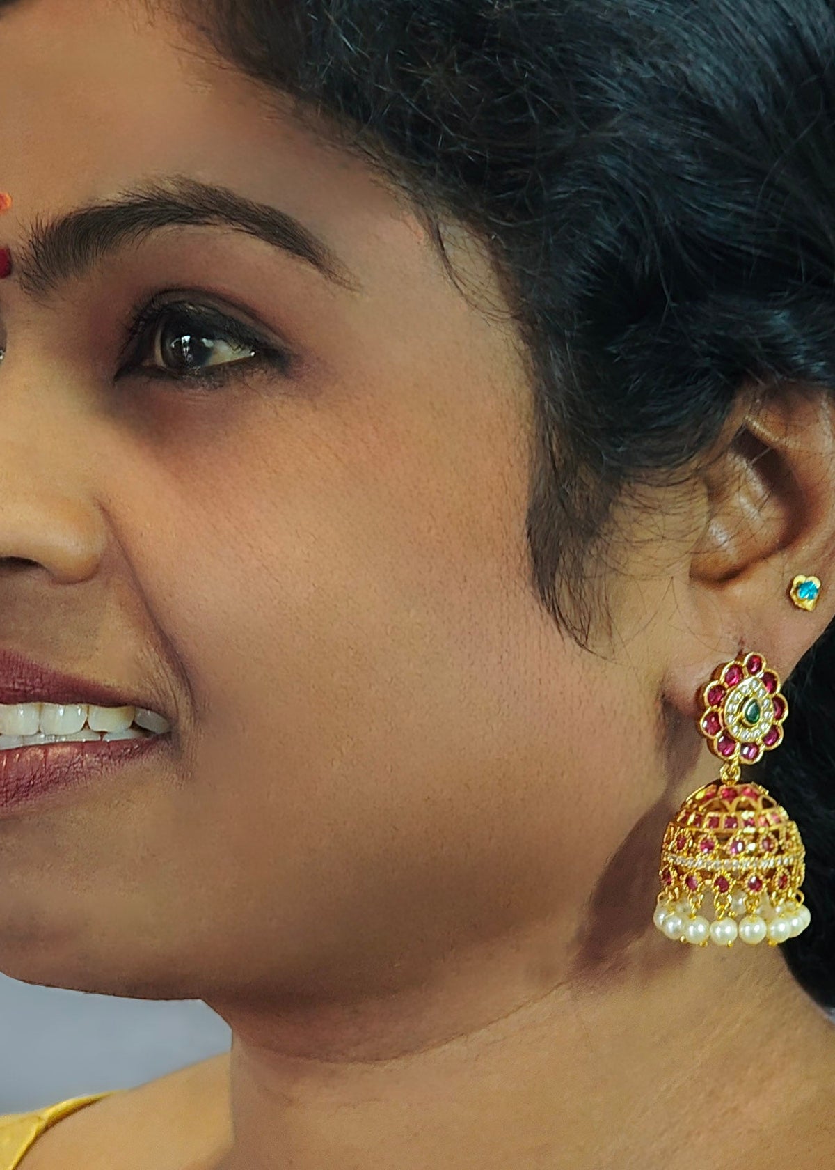 A close-up side profile of a woman wearing a traditional Indian gold jhumka earring with red and green stone accents and white pearl beads.