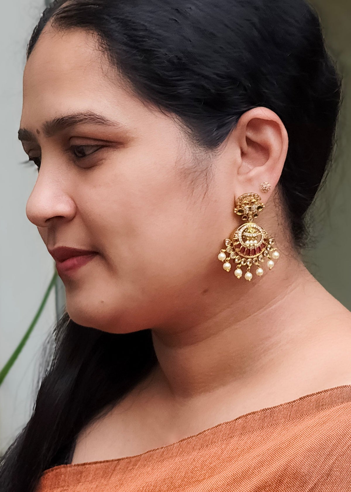 A woman in profile wearing a large, ornate gold-toned traditional Indian jhumka earring with red and white stone detailing and dangling pearl-like beads.