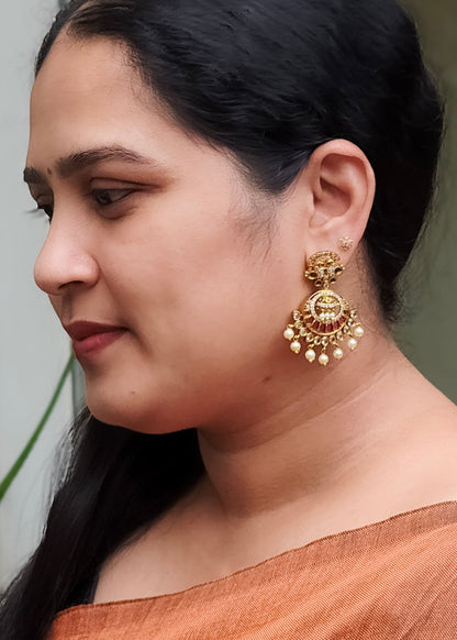 A woman in profile wearing a large, ornate gold-toned traditional Indian jhumka earring with red and white stone detailing and dangling pearl-like beads.