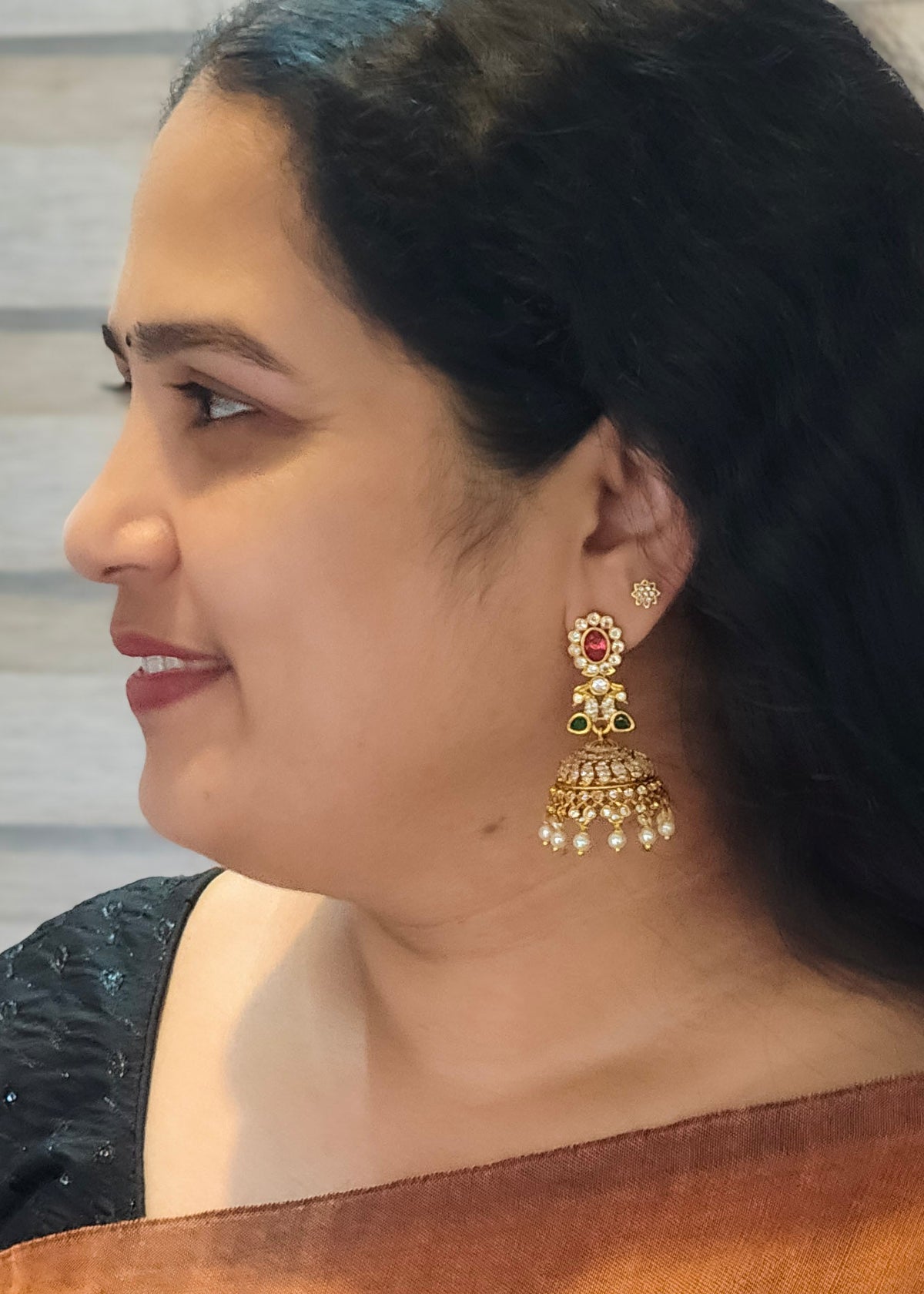 A woman in profile wearing ornate gold-toned jhumka earrings with red and green stone accents and white pearl-like drops.