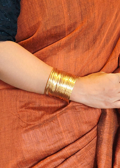A close-up of a person's forearm and hand wearing a wide, multi-strand gold bangle, draped in an orange-brown fabric.
