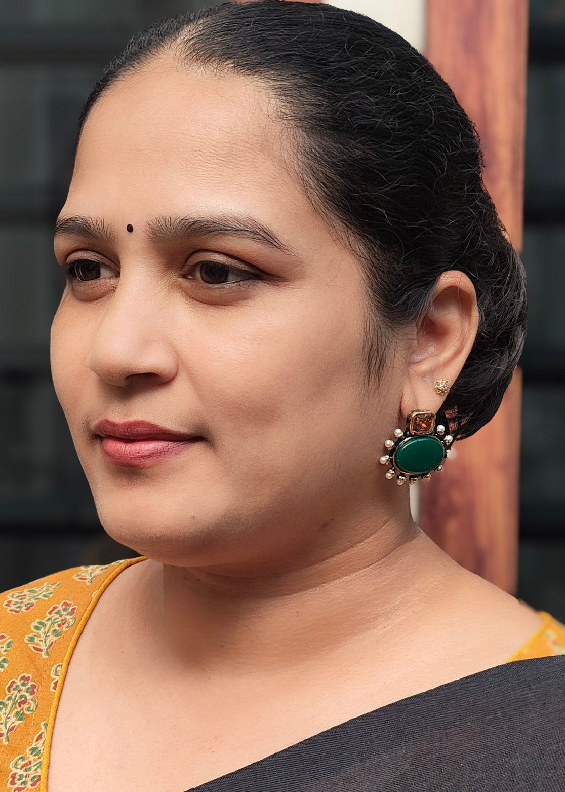 A woman with dark hair tied back, wearing  a green teardrop earring, shown in a close-up portrait.