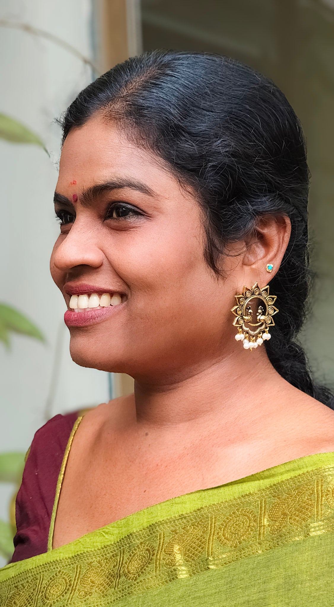 A smiling woman in profile wearing a green saree with gold zari detailing and a maroon blouse, featuring a prominent gold earring.