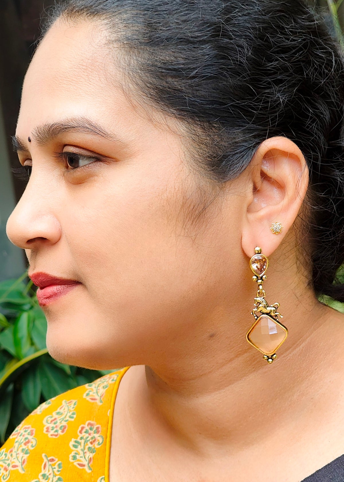 A close-up side profile of a woman wearing a gold-toned dangle earring with a peach-colored stone, alongside a matching small stud earring.