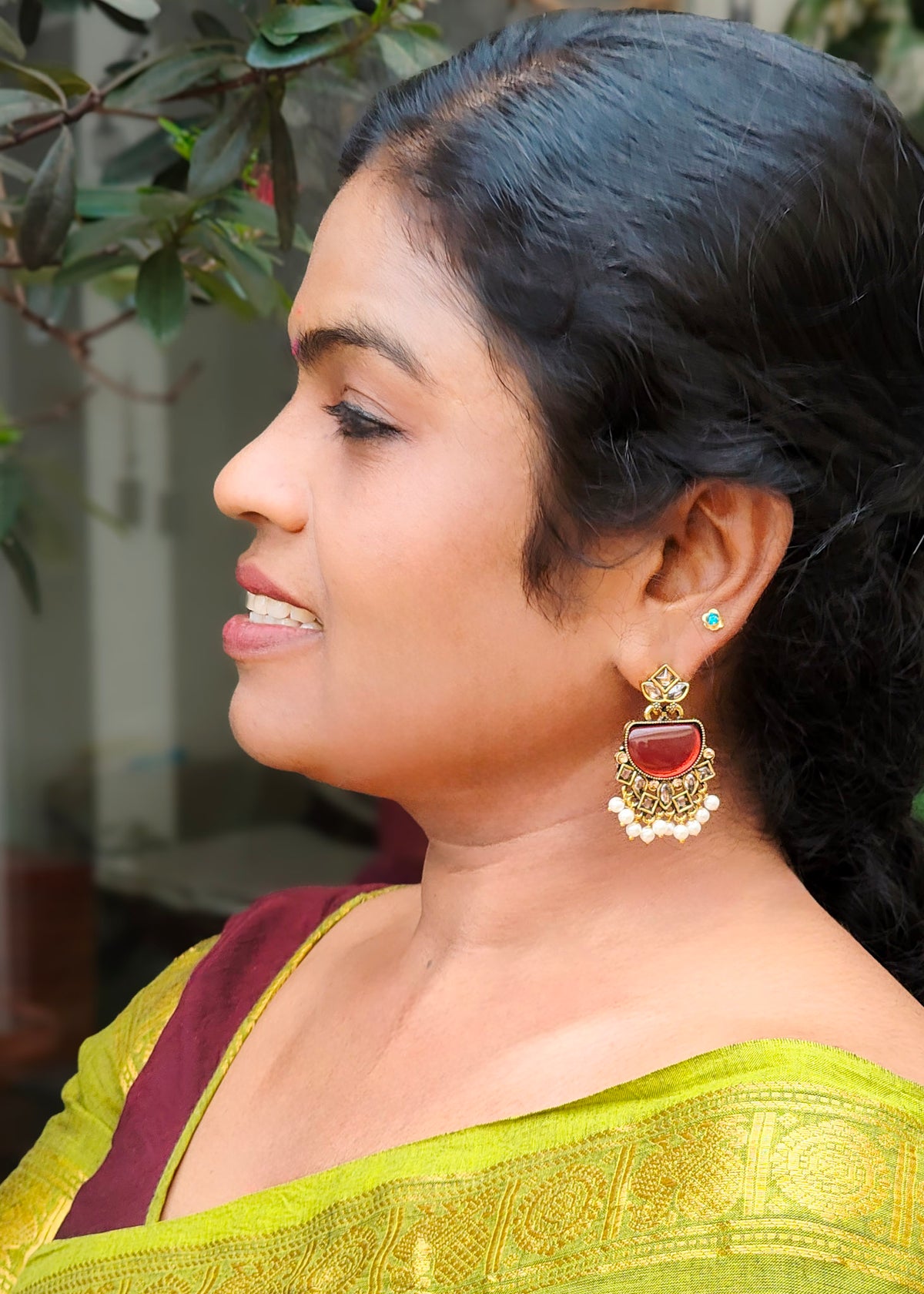 A close-up side profile of a woman wearing a gold-toned traditional Indian jhumka earring with a red stone and white bead border.