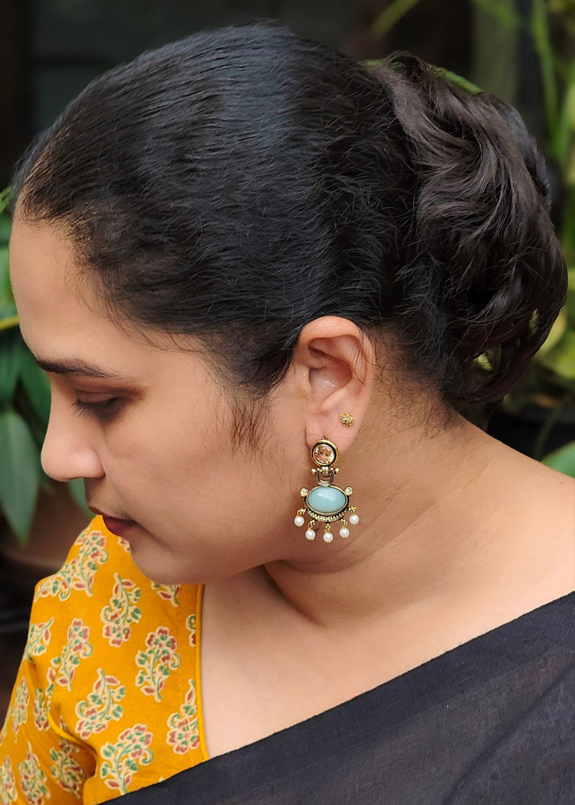 A woman in profile wearing a prominent gold-toned earring with a light blue stone and white pearl accents.