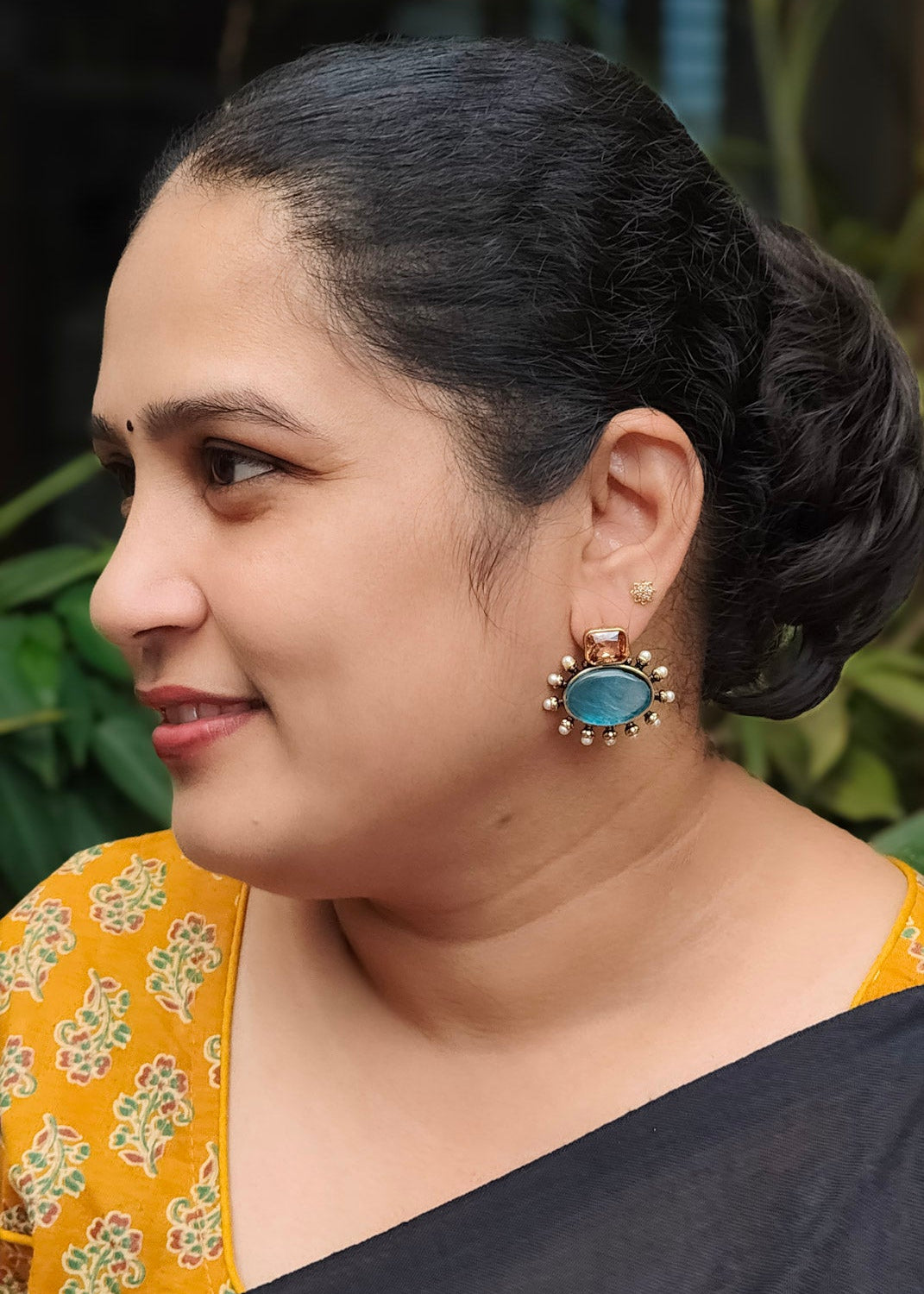 A woman in profile wearing a black saree with a prominent gold-toned earring with a blue stone, 