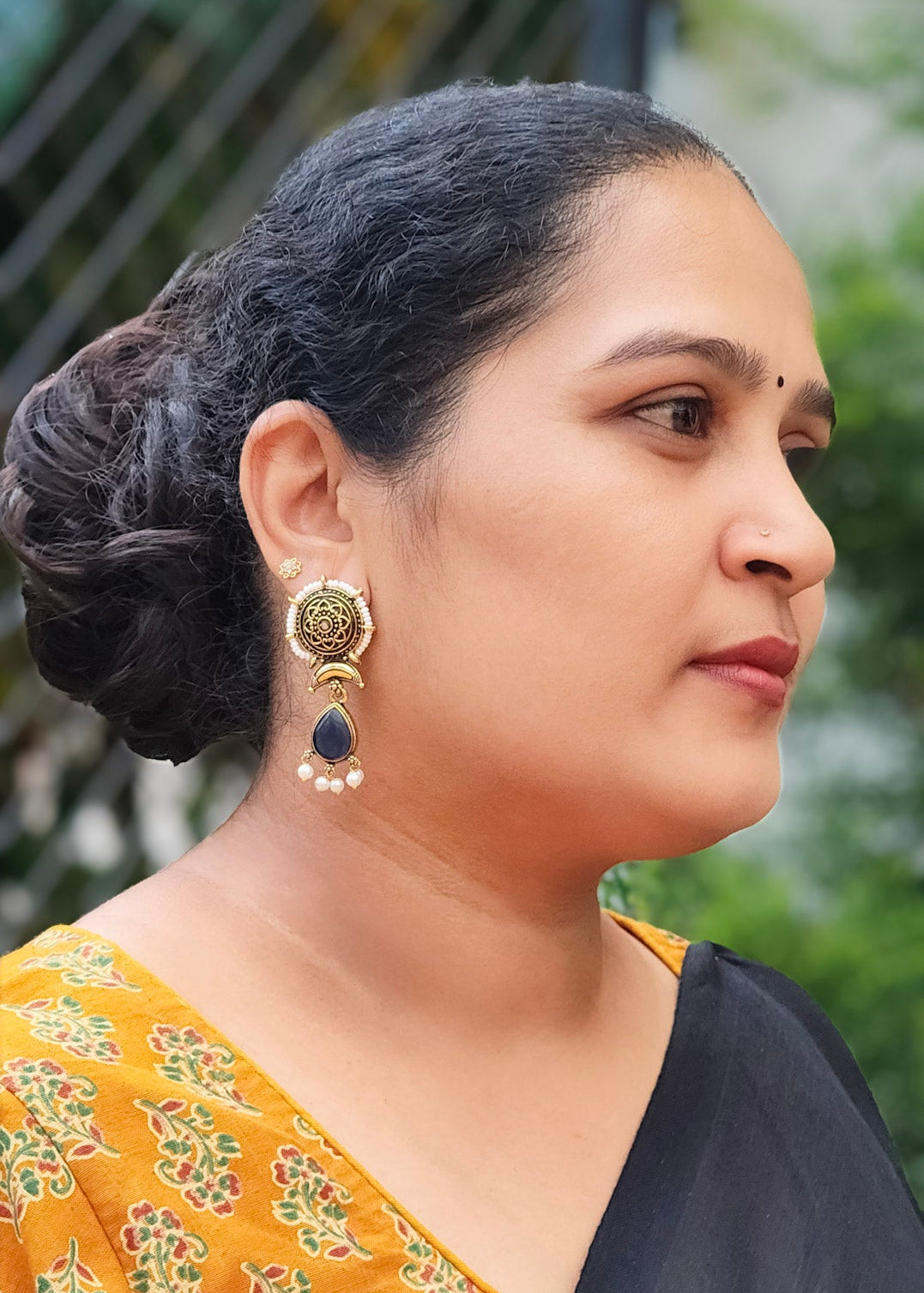 A woman in profile wearing a black saree with a mustard yellow floral printed blouse and a gold-toned earring with a dark blue stone.