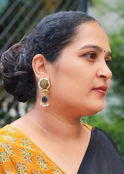 A woman in profile wearing a black saree with a mustard yellow floral printed blouse and a gold-toned earring with a dark blue stone.
