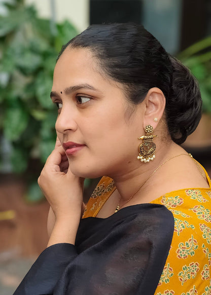 A woman in profile wearing a black dupatta and a mustard yellow printed blouse with gold earrings, set against a soft-focus background of green foliage.