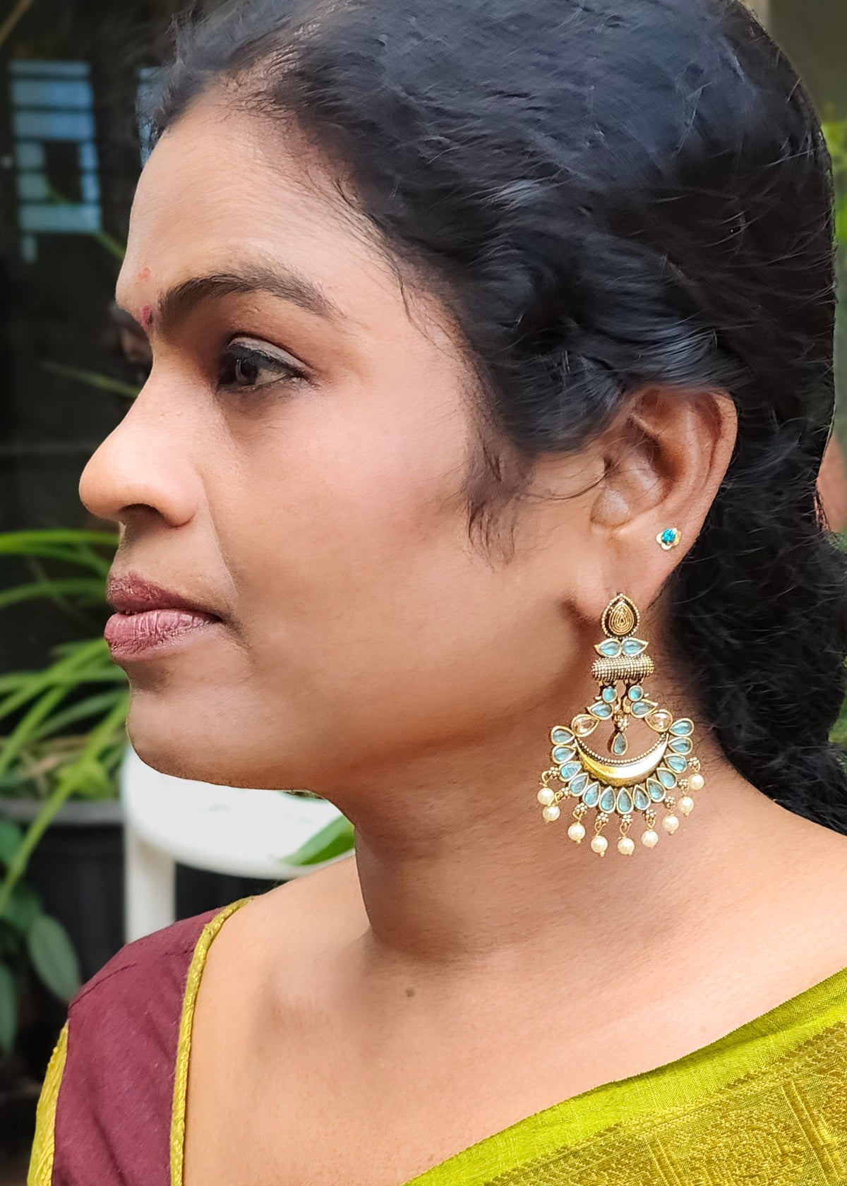 A woman in profile wearing a large gold-toned traditional jhumka earring with light blue stones and white pearl-like dangling beads, 