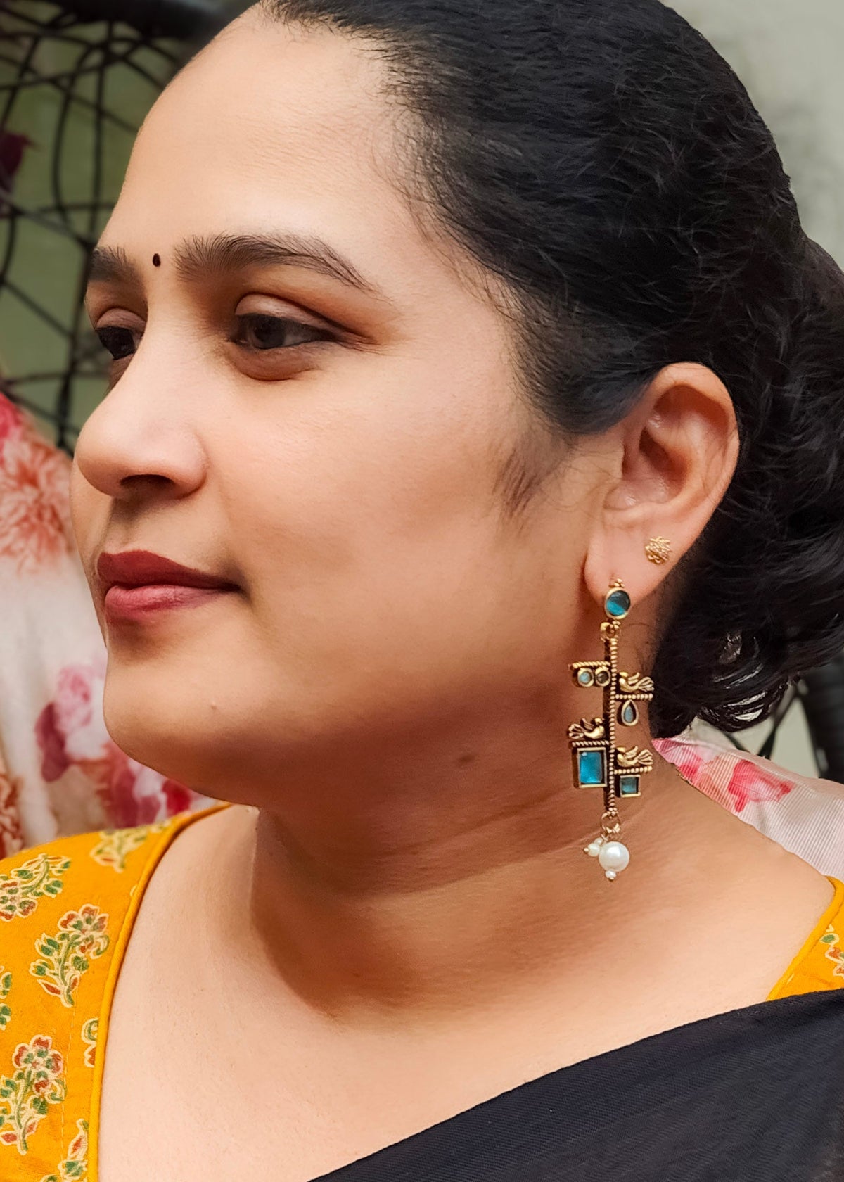 A close-up side profile of a woman wearing a yellow and black saree with ornate gold-toned dangle earrings featuring blue and white stones.