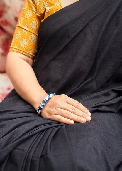 A close-up of a person wearing a black sari with a mustard yellow patterned blouse, resting their hand on their lap while wearing a blue beaded bracelet.