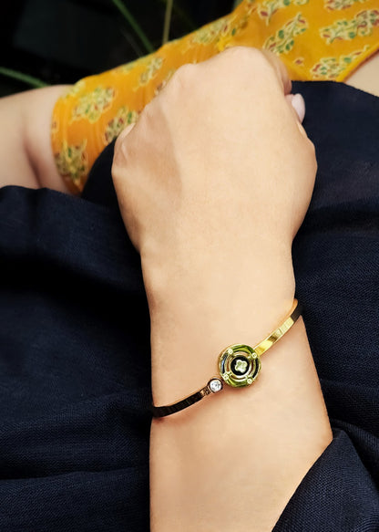 A close-up of a person's wrist wearing a gold and green floral bangle bracelet with a small clear stone accent, set against dark navy clothing and a yellow patterned fabric.