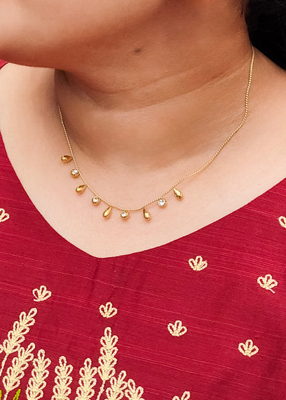 A close-up of a person wearing a delicate gold necklace with small teardrop-shaped gold beads and a few clear stones, paired with a deep red embroidered top.
