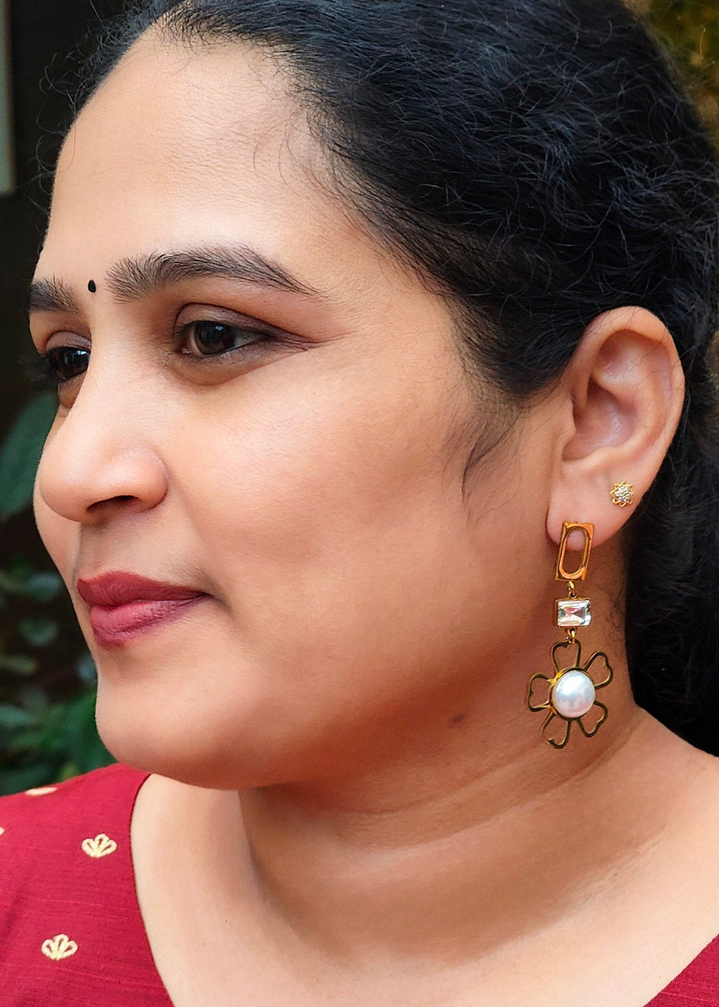 A woman wearing a red embroidered top and a gold-toned dangle earring with a white pearl accent.