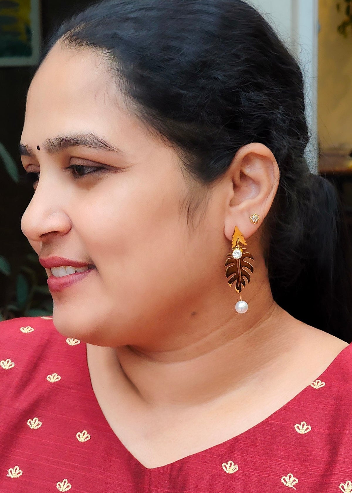 A woman wearing a gold-toned leaf-shaped dangle earring with a white pearl drop, paired with a red patterned top.