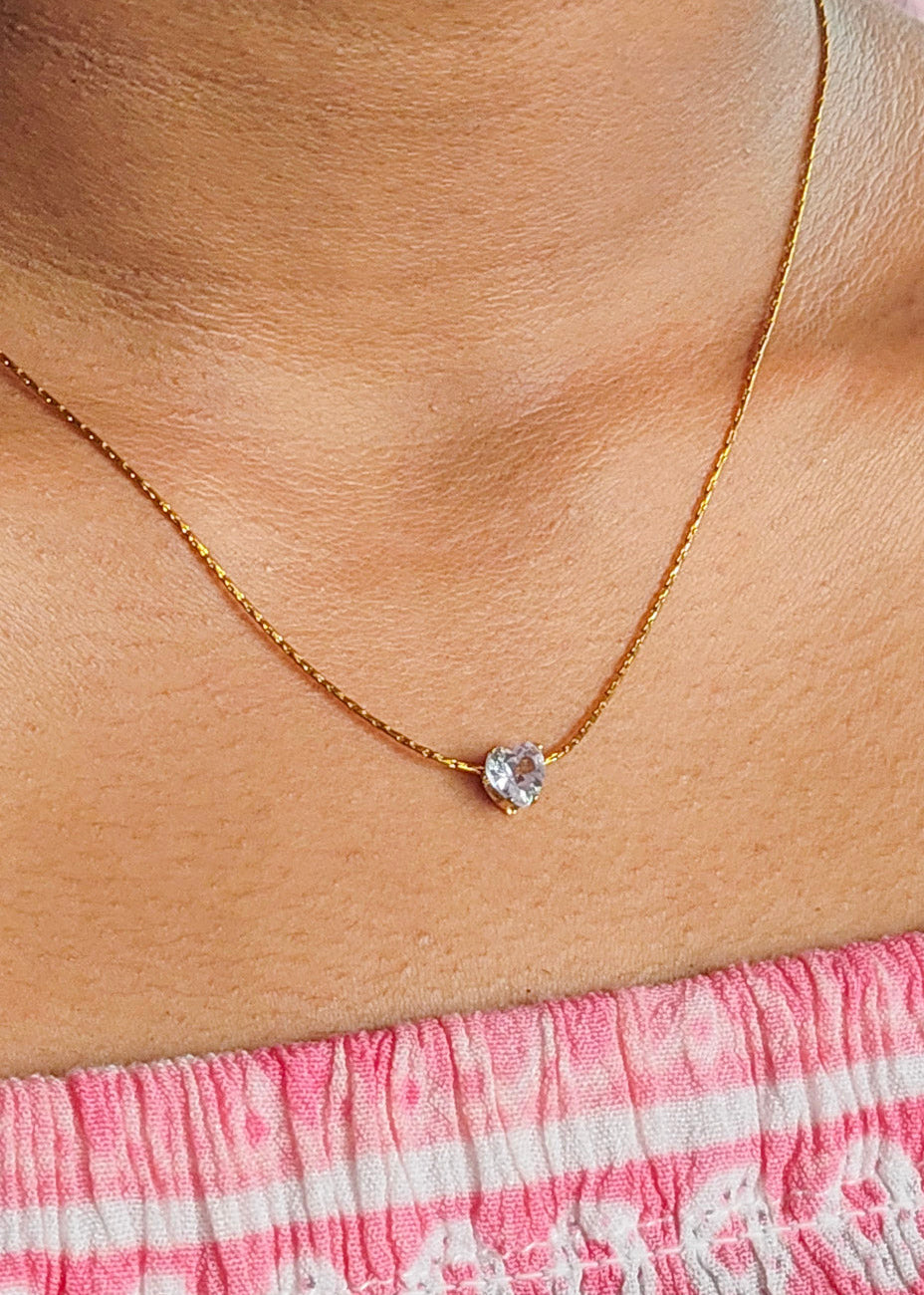 A close-up of a person's neck and upper chest wearing a delicate gold chain necklace with a small clear heart-shaped gemstone pendant, paired with a pink patterned top.