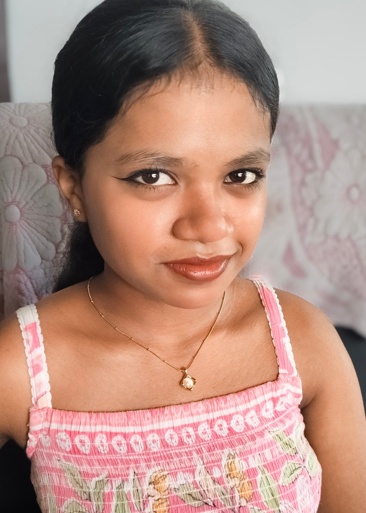 A young girl with dark hair pulled back, wearing a pink patterned spaghetti-strap top and a delicate gold necklace with a small flower-shaped pendant, looking directly at the camera.