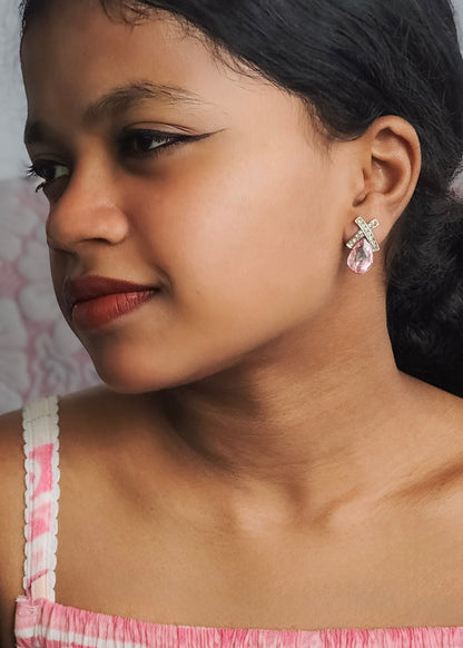 A close-up portrait of a young woman wearing a dangling earring with a silver-tone bow and clear faceted gemstone. The background is a soft, out-of-focus light pink and white patterned wall.