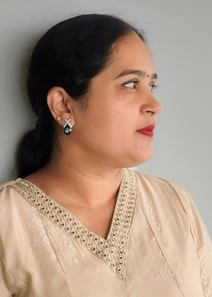 A woman in profile wearing a beige embroidered top and a silver-toned teardrop earring with a dark blue stone, photographed against a plain light gray wall.