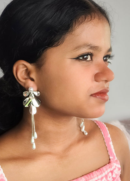 A close-up side profile of a young girl wearing a pair of long, dangling earrings with a silver-toned bow design, green leaf accents, and white pearl-like drops.