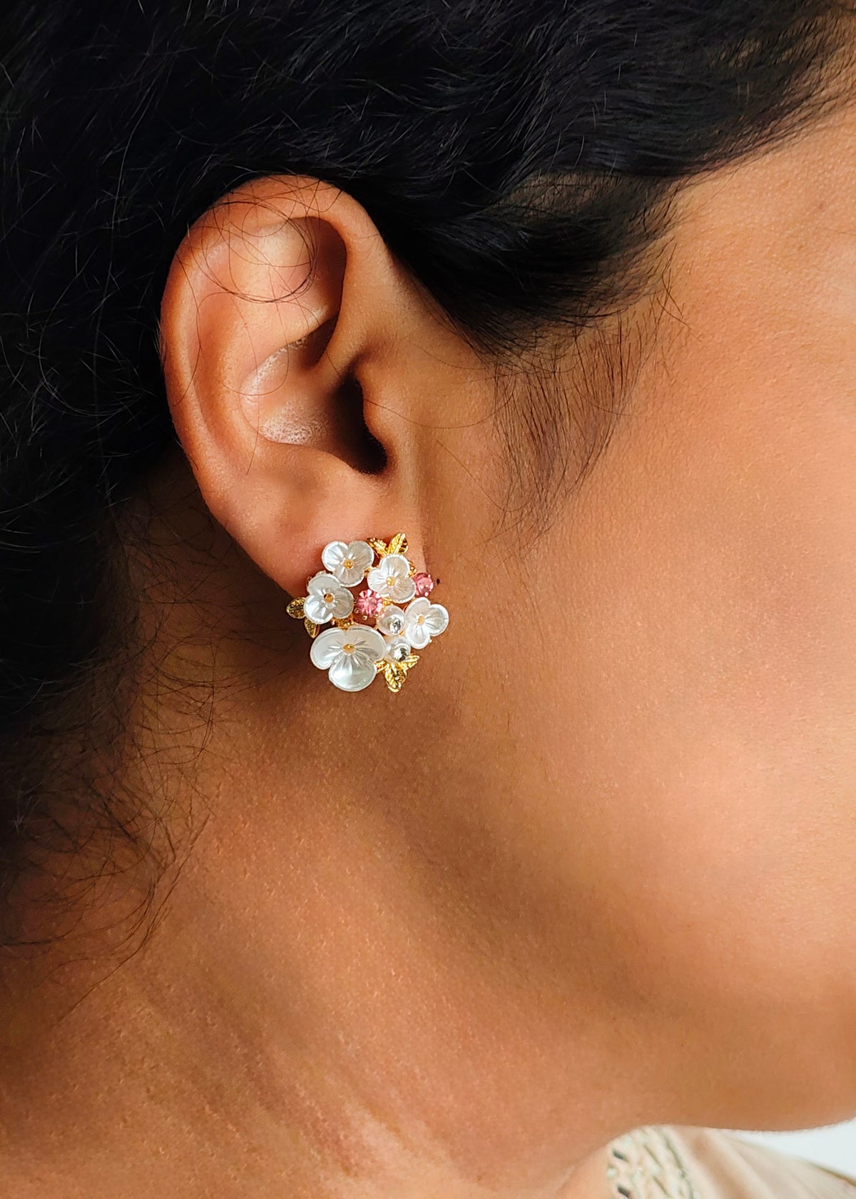 A close-up of a person's ear wearing a small floral stud earring with white flower petals, gold-toned metal, and tiny pink accents.