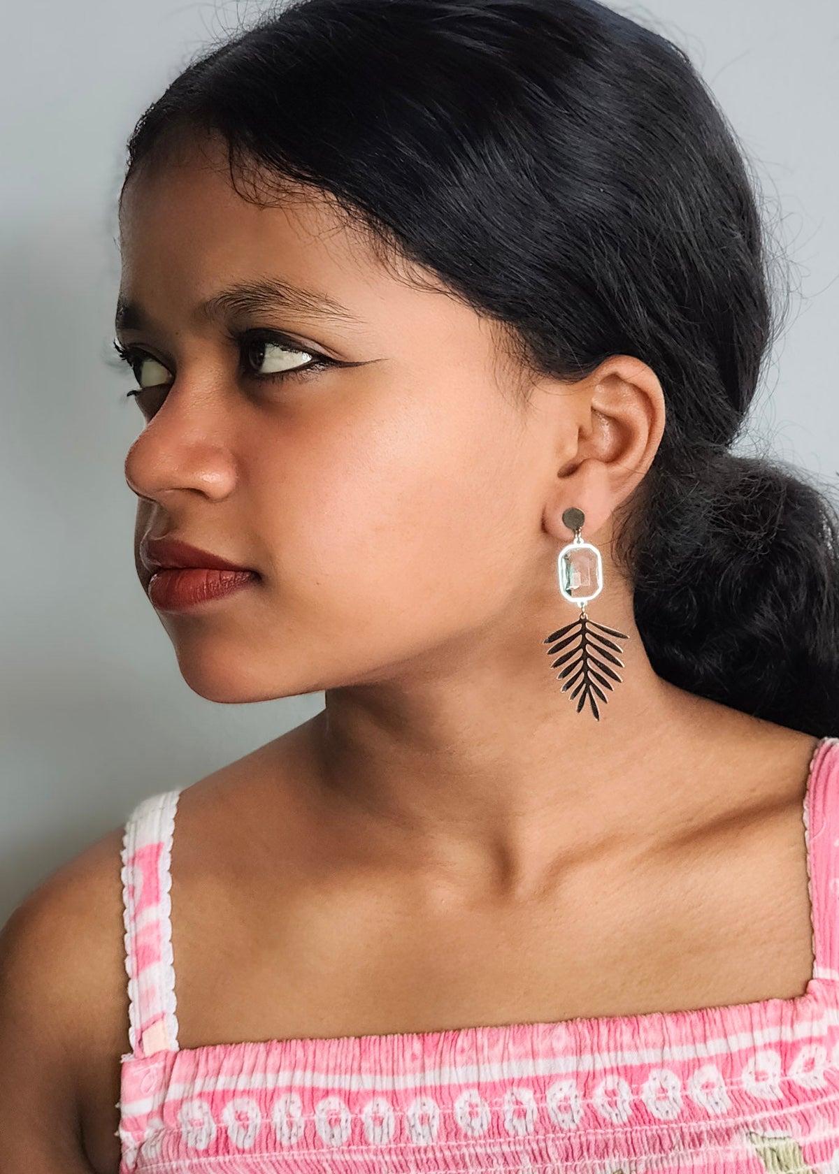 A close-up side profile of a young girl wearing a silver-toned dangle earring with a black feather-like drop and a clear rectangular stone.