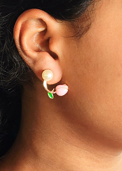 A close-up of a person's ear wearing a small curved earring with a white pearl-like top, a pink flower-shaped drop, and a green leaf accent.