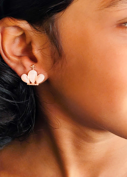 A close-up of a model's ear wearing a small gold-tone stud earring with three pale pink petal-like drops and tiny clear stones along the bottom.