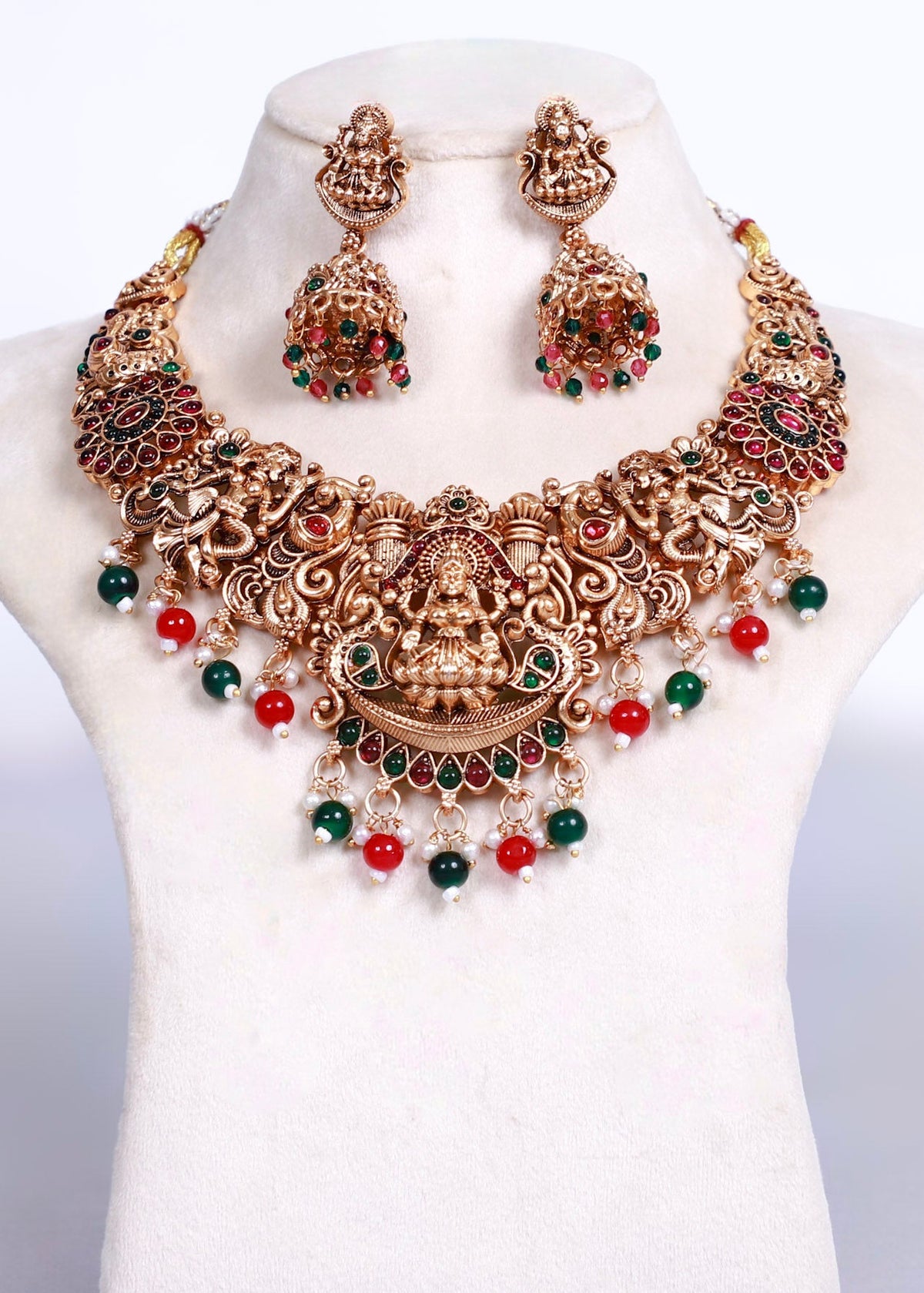 An ornate traditional Indian jewelry set featuring a statement necklace and matching earrings displayed on a white mannequin bust. The jewelry is made of gold-toned metal with intricate filigree work, decorated with red and green stones and small red and green bead drops.