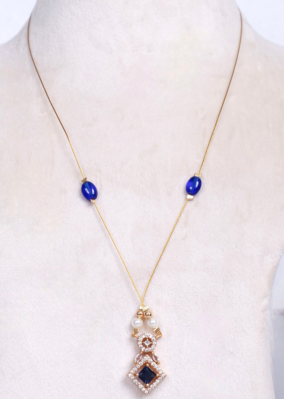 A gold necklace displayed on a cream-colored mannequin bust, featuring two blue oval beads on the chain and a long drop pendant with white pearls, a small rose gold floral accent, and a central dark blue square stone surrounded by clear rhinestones.