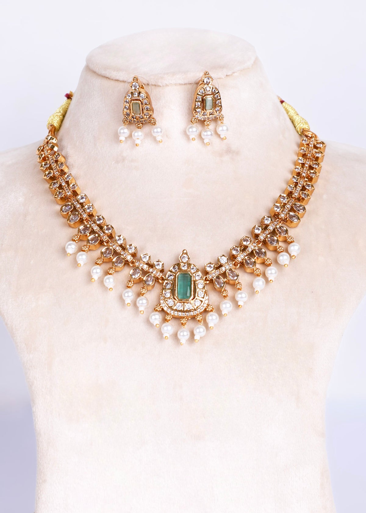 Gold-toned traditional Indian jewelry set featuring a statement necklace and matching earrings, embellished with green stones, clear crystals, and white faux pearls, displayed on a cream-colored mannequin bust.