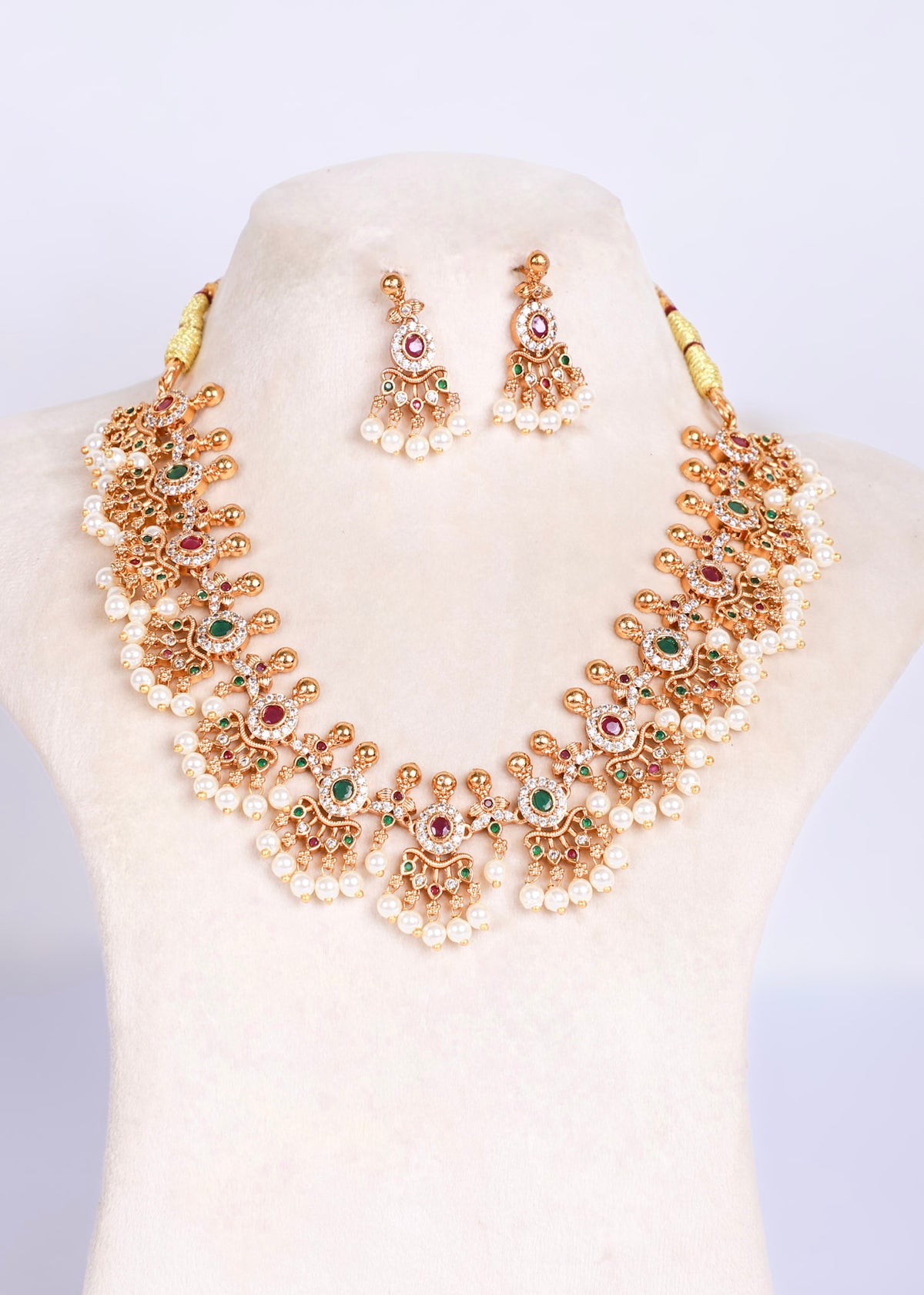 An ornate gold-toned statement necklace with matching drop earrings, displayed on a cream-colored jewelry bust. The jewelry features intricate filigree detailing, white faux pearls, and small red and green gemstone accents.